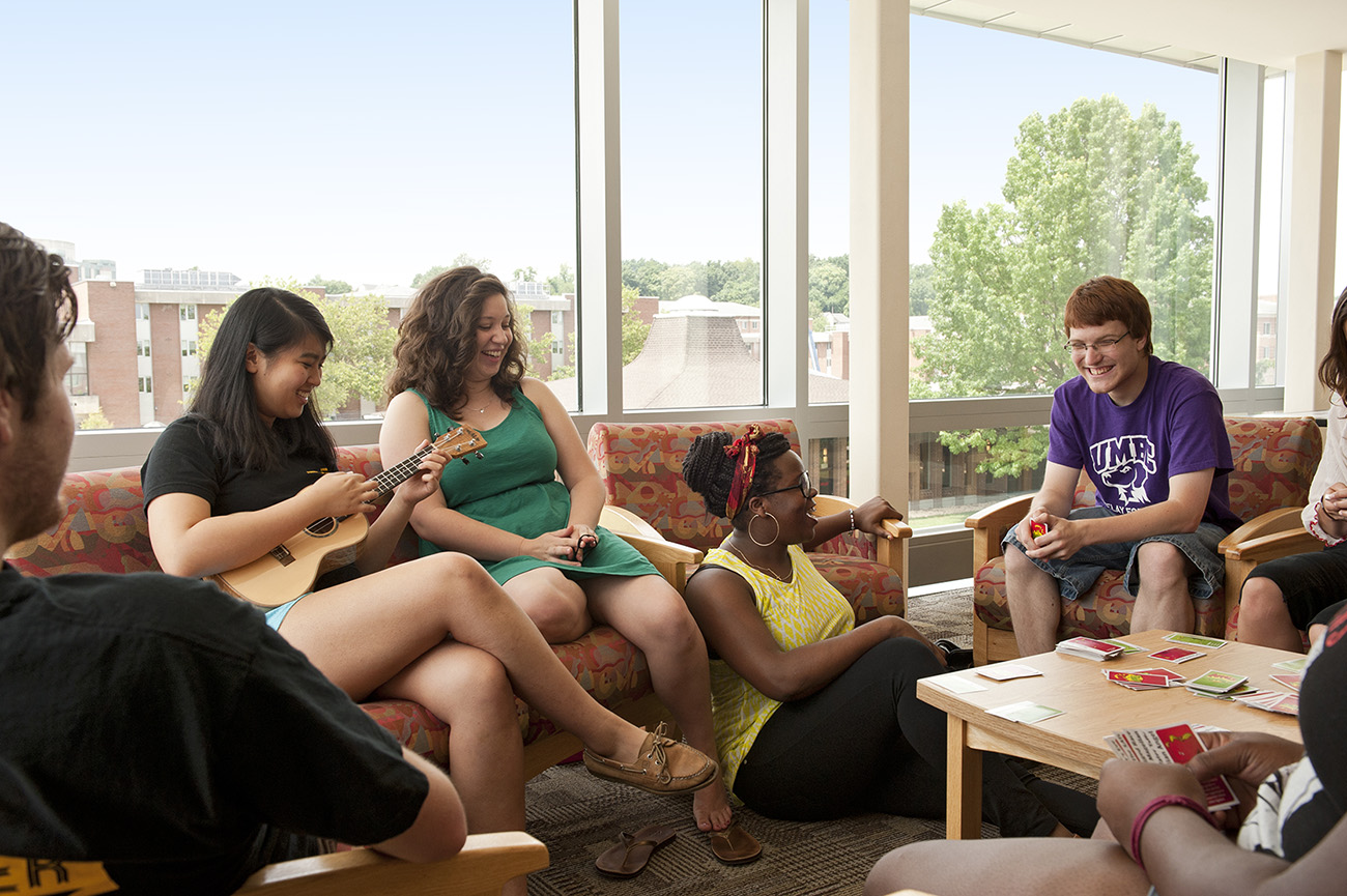 Six diverse students relaxing on couches with games and a guitar, and a panoramic view of the lush campus.