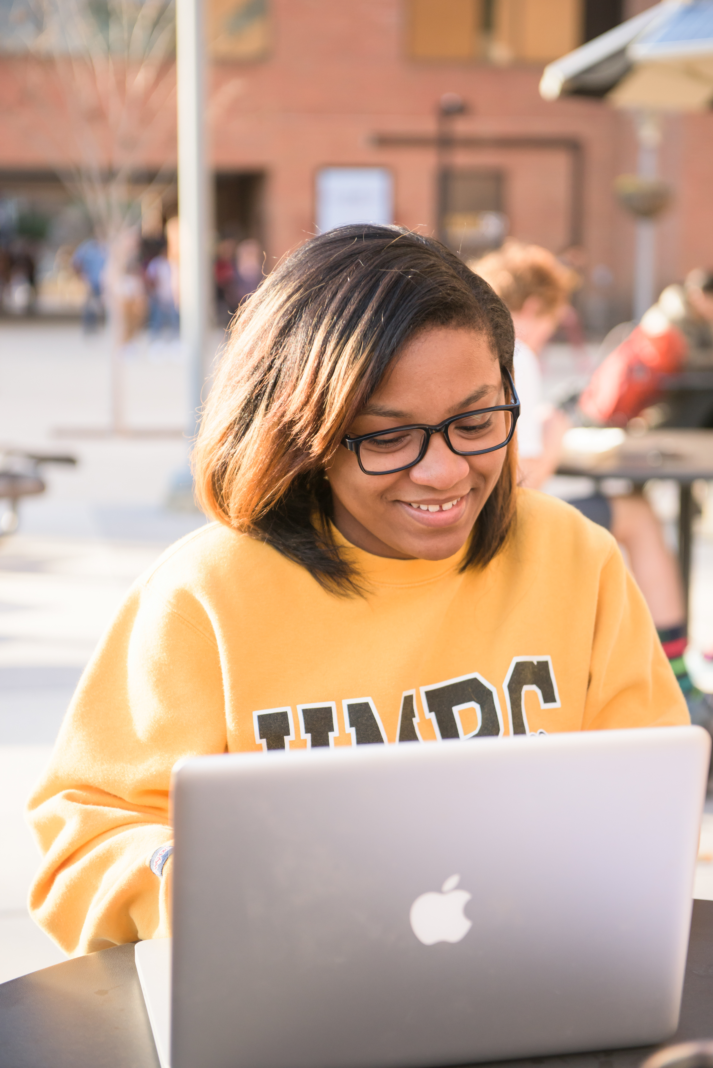 A student wearing a UMBC hoodie sitting outdoors working happily on her computer.